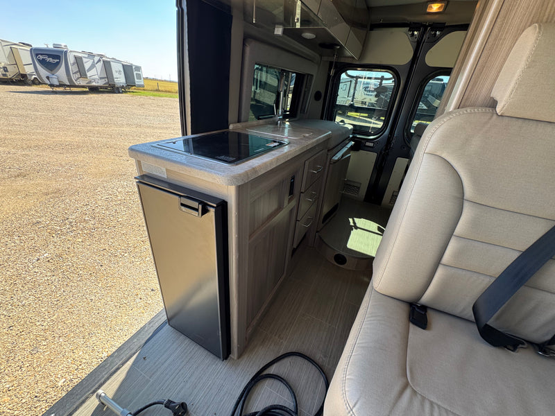 Explore this RV rental's compact interior, featuring a kitchen with a built-in stovetop, sink, and refrigerator, beside a comfortable passenger seat. — photo 10