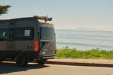RV rental ready: A grey camper van with all-terrain tires parked on a sunny coastal road, ocean view & distant rigs.