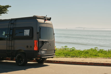 RV rental ready: A grey camper van with all-terrain tires parked on a sunny coastal road, ocean view & distant rigs.