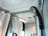 Adjustable black showerhead in a compact RV rental bathroom, with light privacy curtains and reflective surfaces.
