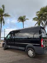 Sleek black RV rental van reflecting palm trees and cloudy sky by a tropical waterfront. Ideal for your next journey.