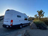 This white RV rental camper van is parked amidst Joshua trees and boulders in a sunny desert landscape, perfect for adventure.