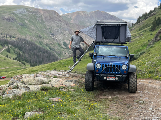 Man sets up a blue Jeep with rooftop tent on a mountain trail, perfect for an adventurous rv rental experience.