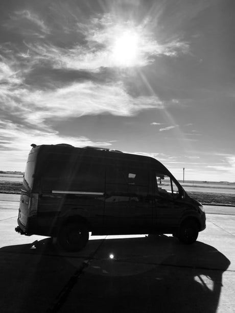 Monochrome image of a dark RV rental van parked on concrete, silhouetted against a sunny, clouded sky.