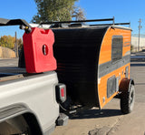 A silver pickup truck tows an orange and black teardrop camper, equipped with a roof rack and gas can. Great RV rental option!