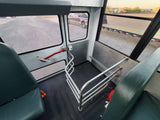 Inside an RV rental, showing green seats, a red emergency exit, and a metal safety railing. Gravel lot visible outside.