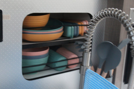 A compact RV rental kitchen featuring colorful stacked dishes (bowls/plates) and cooking utensils next to a modern coiled faucet.