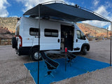Scenic rv rental setup with a white camper van, awning, table, and chairs on a gravel lot with red rocks in the background.