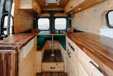 Cozy rv rental camper van interior. Features include wood paneling, a kitchenette, dinette with green cushions, and ocean views.