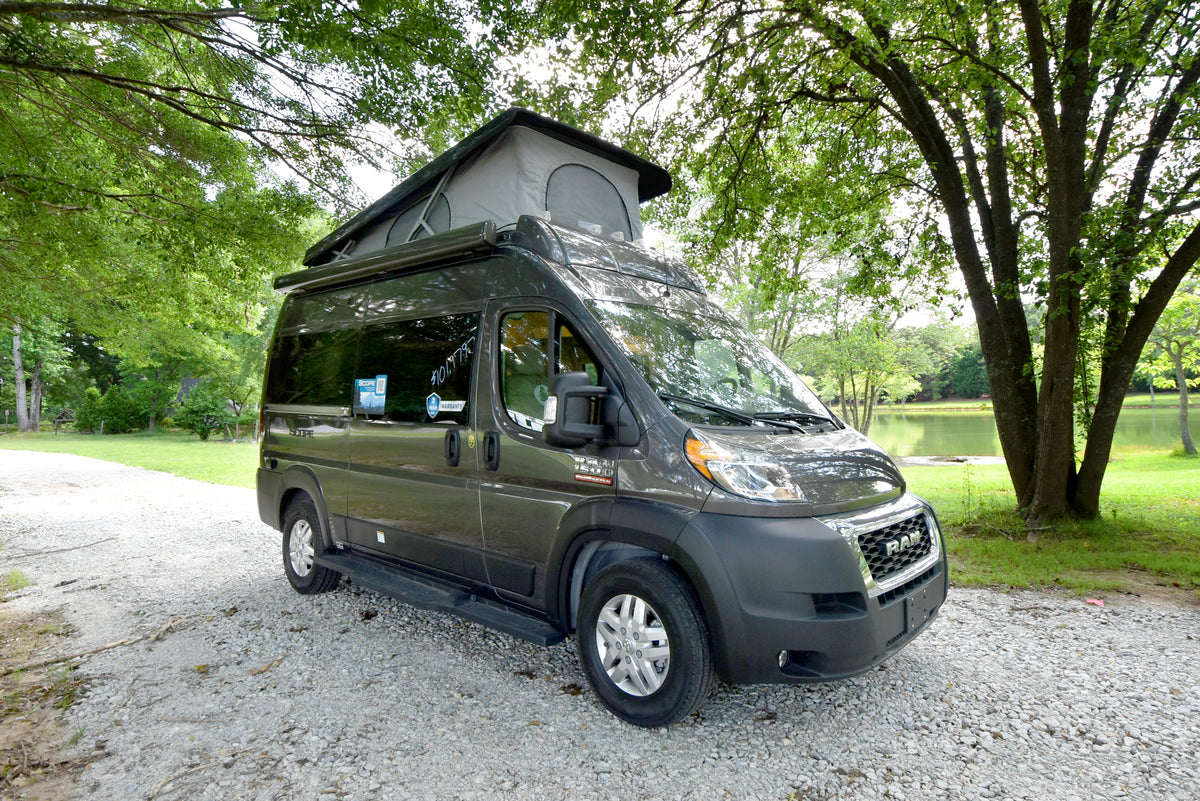 Modern dark grey RV rental camper van with pop-top roof on a gravel path, amidst green trees and a serene lake.