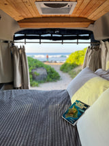 Relaxing inside an RV rental. A Hawaii guidebook rests on the bed, with a serene beach view beyond.