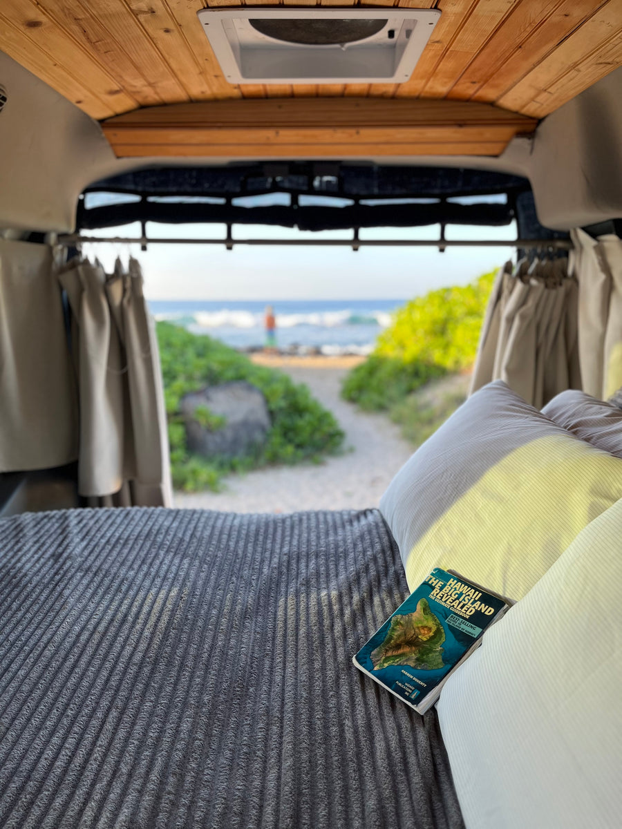 Relaxing inside an RV rental. A Hawaii guidebook rests on the bed, with a serene beach view beyond.