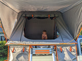 A happy toddler peeks from a grey rooftop tent mounted on a vehicle, accessible by a ladder. Perfect for RV rental trips!