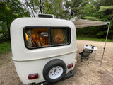 RV rental: white camper with awning, table, chair. An adult and two kids are cozy inside. Perfect for outdoor family camping.