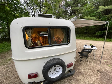 RV rental: white camper with awning, table, chair. An adult and two kids are cozy inside. Perfect for outdoor family camping.