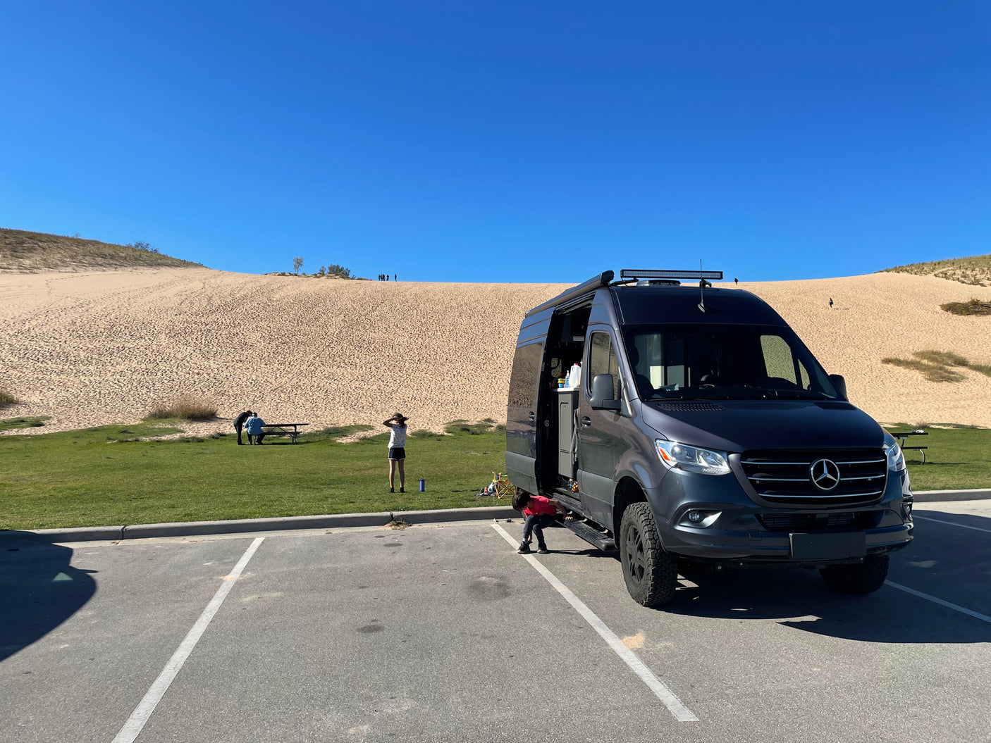 RV rental: Dark grey Mercedes Sprinter van parked by large sand dunes. People enjoy this sunny outdoor scene, perhaps a camping trip.