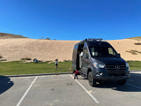 RV rental: Dark grey Mercedes Sprinter van parked by large sand dunes. People enjoy this sunny outdoor scene, perhaps a camping trip.