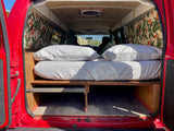 Open back of an RV rental showing a comfortable bed with white linens, built-in storage, and tropical wall decor.
