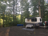 Enjoying an rv rental at a scenic wooded campsite. White/blue trailer, awning, table, and person setting up by a lake.