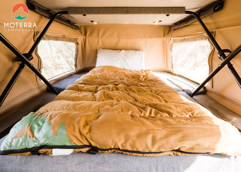 Interior of an RV rental: a cozy bed with a brown sleeping bag (green mountain design) & white pillow, surrounded by bright windows. — photo 5