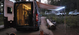 An RV rental camper van illuminated at night, back open revealing its interior, parked under an awning in a forest campsite.