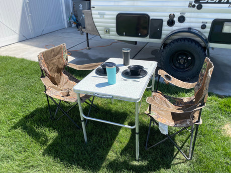Outdoor dining by a small RV rental: two camo chairs, a table with dishes & cups. Ideal for your next camp adventure!