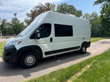 White high-top camper van, a versatile RV rental, parked on an asphalt drive. Features tinted windows & roof setup.