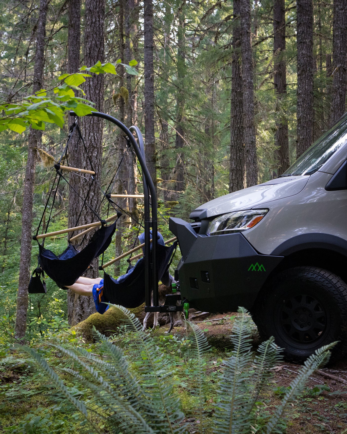 Enjoying the forest in an RV rental! A person relaxes in a hanging chair attached to the van's front bumper, dog peeking out.
