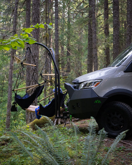 Enjoying the forest in an RV rental! A person relaxes in a hanging chair attached to the van's front bumper, dog peeking out.