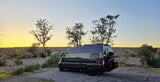 A dark gray camper van, perfect for RV rental, parked at a desert campsite with a beautiful sunset sky.