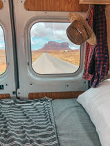 Cozy RV rental with a bed, showing a long desert road through the window leading to a majestic mesa.