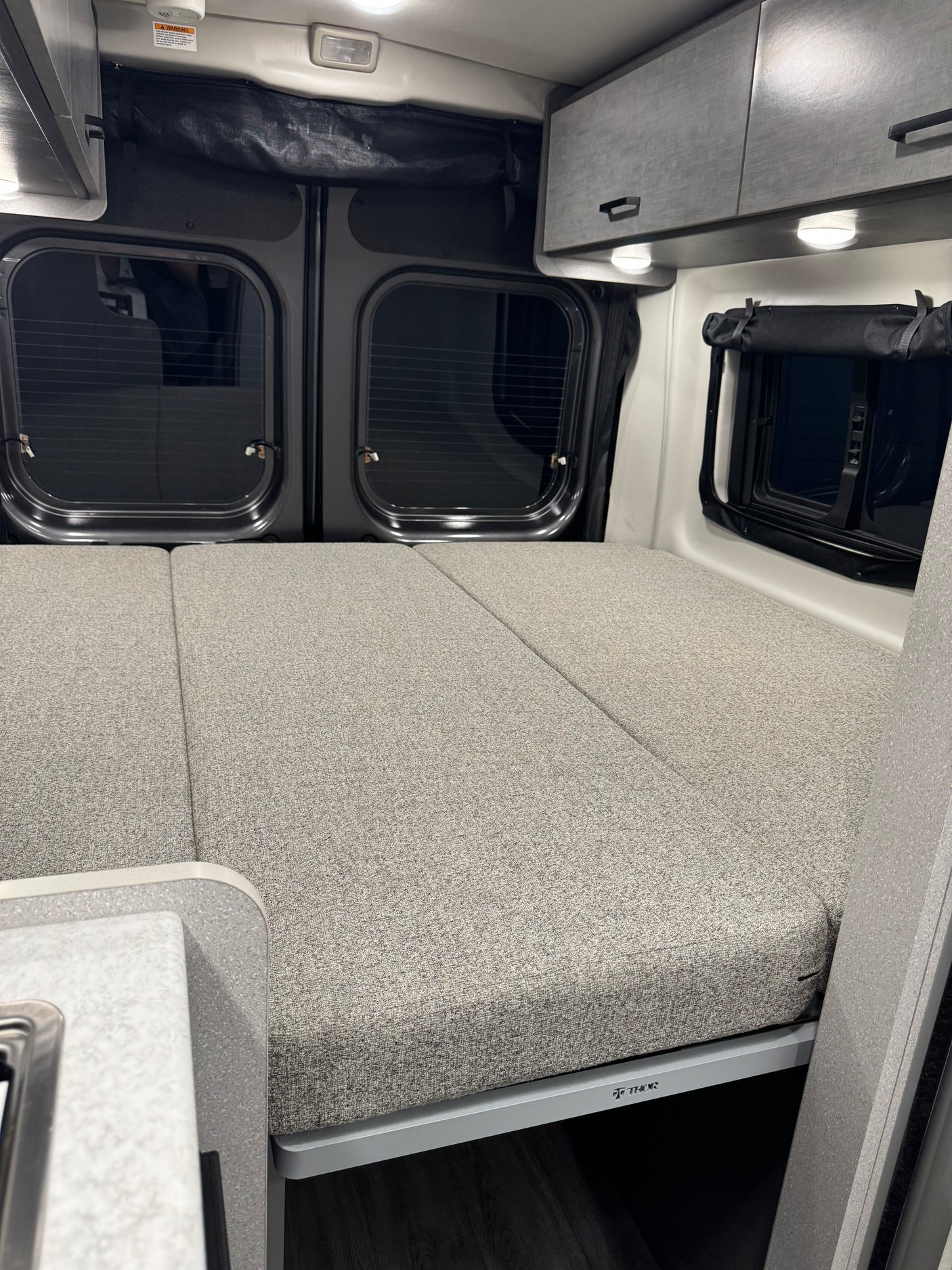 Comfortable RV rental interior with a gray convertible bed, ample overhead storage, and windows, ideal for travel.