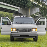 Front view of a white Ford van, ideal for rv rental, with open doors. It's parked on green grass beneath a concrete bridge.
