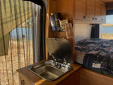 RV rental interior: compact kitchen with sink & supplies, cozy bed. A mesh window reveals a serene lake and wild landscape.