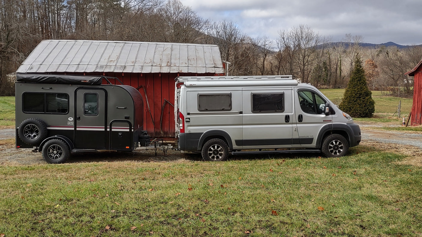 RV rental ready: Silver camper van towing a black off-road trailer by a red barn, ideal for outdoor adventure.
