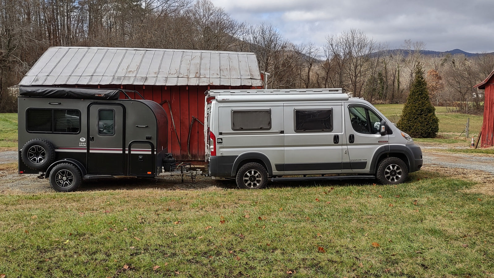 RV rental ready: Silver camper van towing a black off-road trailer by a red barn, ideal for outdoor adventure.