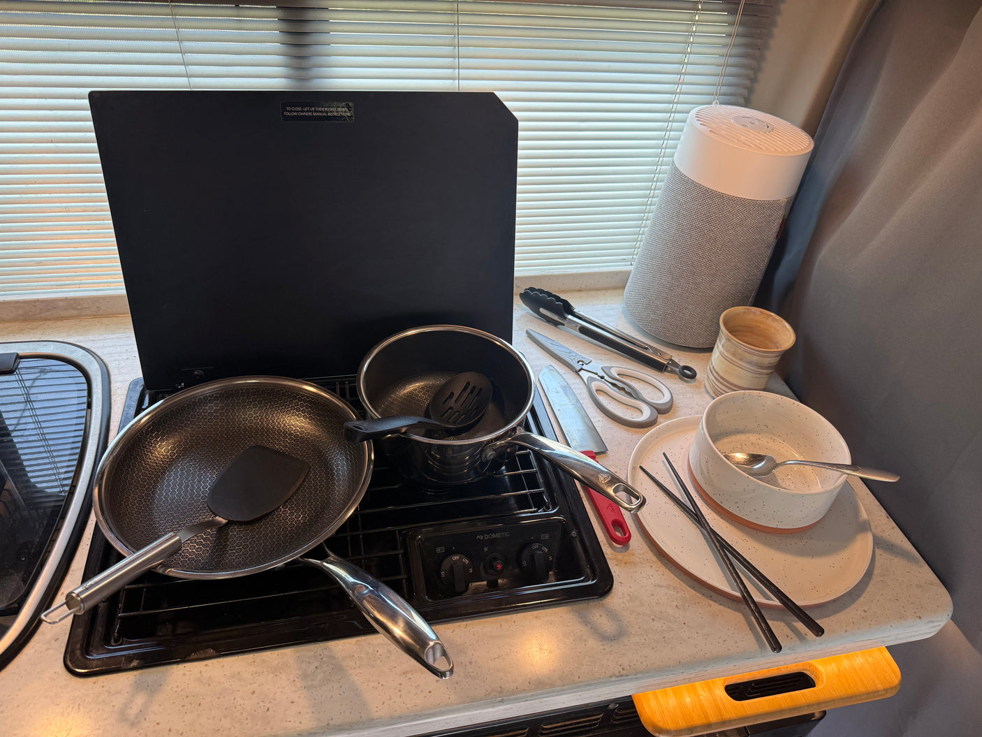 This RV rental kitchen features a Dometic stovetop with a frying pan and pot, plus various cooking utensils and dishes, ready for a meal.