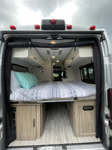 View of an RV rental interior from the rear, showing a bed, overhead storage, and air conditioning for comfortable travel.