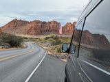 Enjoy a scenic rv rental road trip! Van parked by a winding desert road, with red rock mountains & a large US flag.