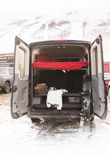 An RV rental with open rear doors reveals a cozy bed, ski gear, and a snowboard, ready for snowy mountain adventures.