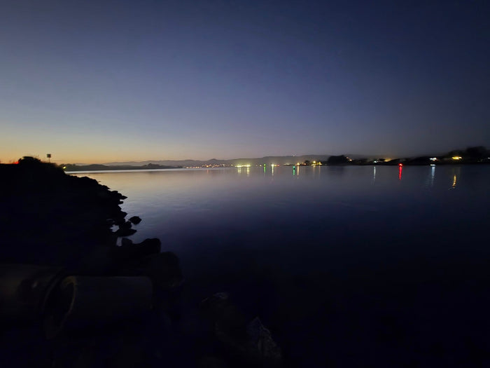 Tranquil water at dusk with glowing distant lights, ideal for an RV rental trip. Dark rocky shore in view.