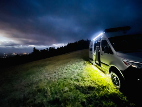 Nighttime view of an RV rental parked on a hill, its bright lights illuminating the grassy ground under a dark, cloudy sky with city lights in the distance.
