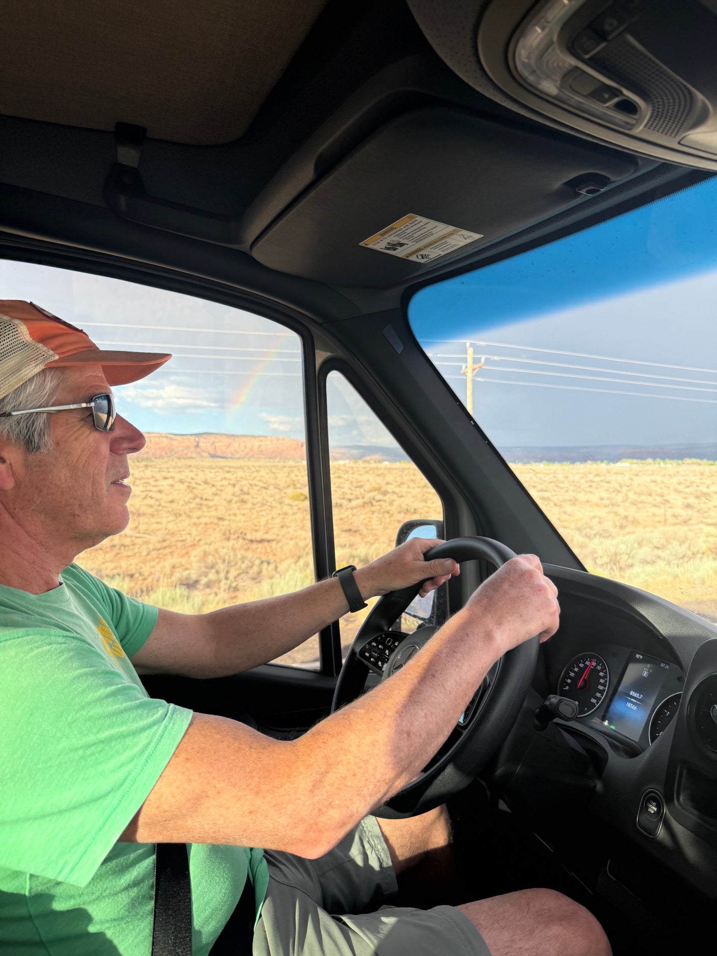 RV rental road trip: Driver behind wheel, desert landscape with faint rainbow through window. Dashboard shows 92°F.