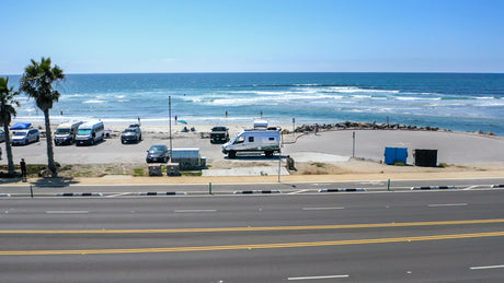 RV rentals parked at a sunny beachside lot. Waves crash as people enjoy the ocean view and sand.