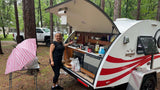 Woman cooking in an outdoor kitchen of a Boondock Lite teardrop RV rental at a scenic campground. Umbrella on table.