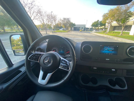 Inside an RV rental van: driver's view showing Mercedes-Benz steering wheel, dashboard display, and outdoor parking lot scene.