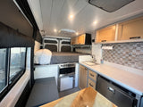 This RV rental interior showcases a cozy bed, compact kitchen with sink & microwave, and a dining area. Ready for your adventure.