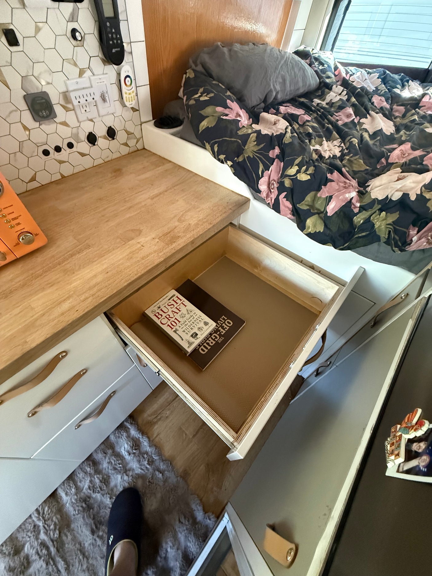RV rental interior: Floral bed, wooden counter, open drawer showing "Bushcraft 101" and "The Living Off-Grid" books.