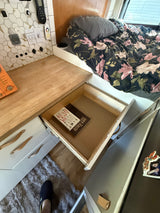 RV rental interior: Floral bed, wooden counter, open drawer showing "Bushcraft 101" and "The Living Off-Grid" books.