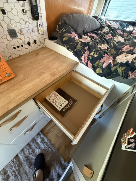 RV rental interior: Floral bed, wooden counter, open drawer showing "Bushcraft 101" and "The Living Off-Grid" books.